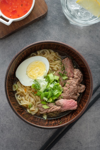 Soupe de ramen asiatique avec bœuf, œuf, ciboulette dans un bol sur fond gris.