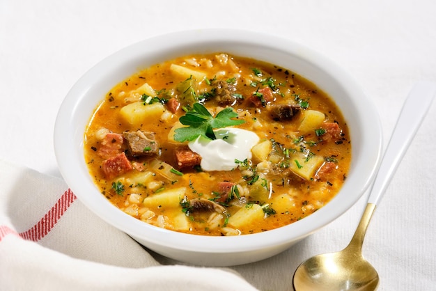 Soupe rassolnik, boeuf, orge, carotte et cornichon. Cuit à base de concombre aigre-doux. Portion de soupe dans un bol blanc sur nappe textile blanc cassé. Servi avec de la crème sure et du persil.