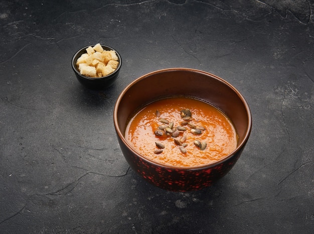 Soupe végétarienne savoureuse à la citrouille dans un bol noir décoré de graines sur fond sombre