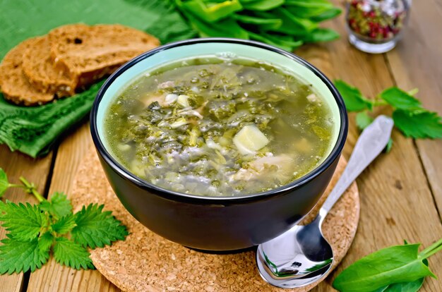 Soupe verte d'oseille, d'ortie et d'épinards dans un bol, cuillère, pain, poivre contre une planche en bois