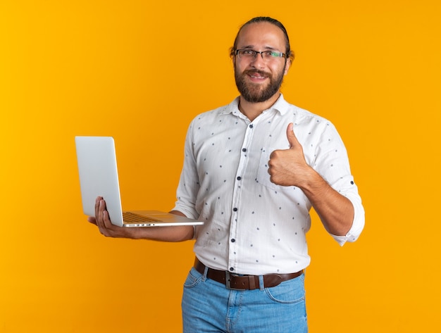 Souriant adulte bel homme portant des lunettes tenant un ordinateur portable regardant la caméra montrant le pouce vers le haut isolé sur un mur orange