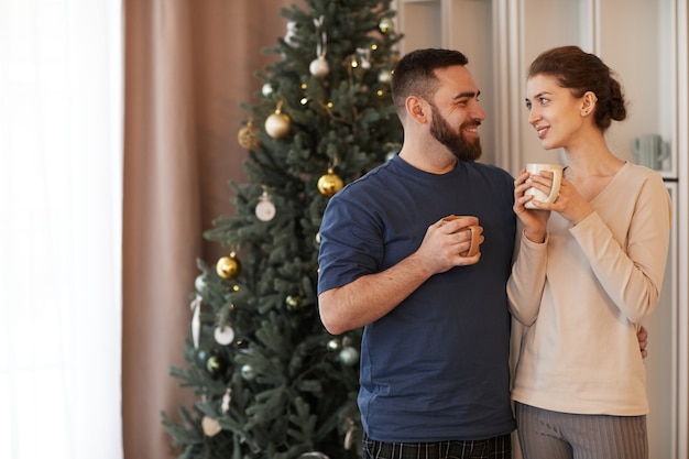 Souriant aimant jeune couple caucasien debout contre l'arbre de Noël et embrassant tout en buvant c...