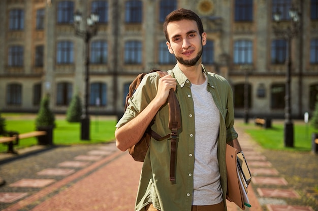 Souriant beau étudiant aux cheveux noirs avec un sac à dos posant pour la caméra contre le bâtiment de l'université