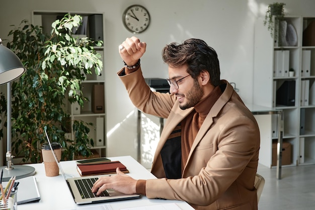 Souriant beau gestionnaire du Moyen-Orient dans des lunettes faisant un geste oui et utilisant un ordinateur portable tout en obtenant un résultat