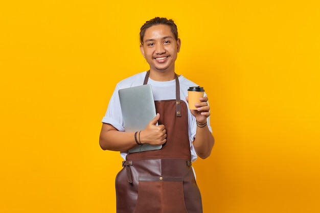 Souriant beau jeune homme tenant un ordinateur portable et tenant une tasse de café sur fond jaune