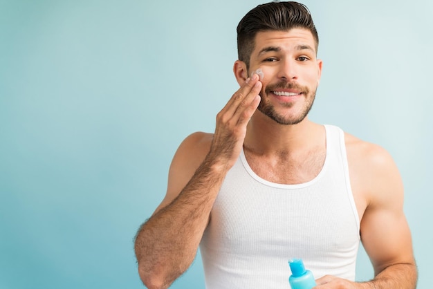 Souriant bel homme appliquant une crème anti-âge sur la joue tout en établissant un contact visuel sur fond uni