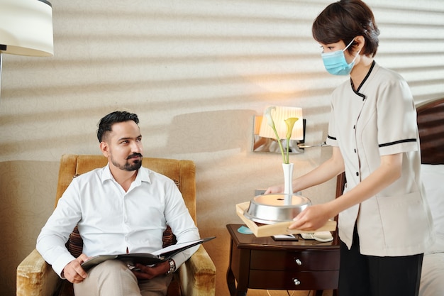 Souriant bel homme mûr regardant la femme de chambre dans un masque médical apportant un petit-déjeuner frais à la chambre d'hôtel