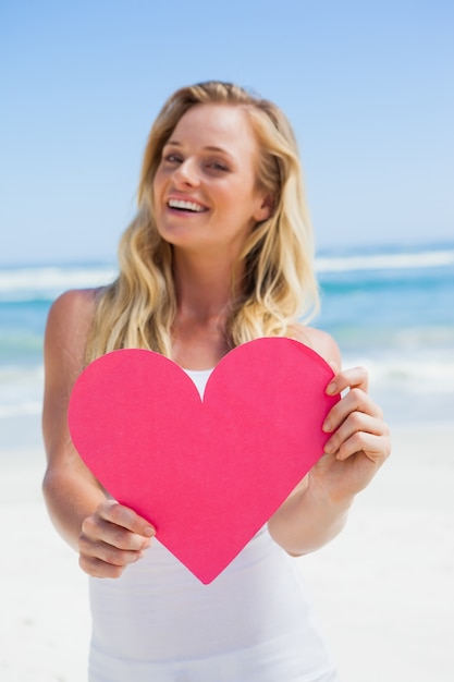 Souriant blonde montrant un coeur rose sur la plage