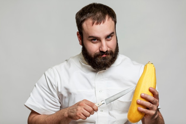 Souriant chef masculin barbu avec un couteau et des courgettes jaunes dans ses mains. Un professionnel en veste blanche s'apprête à cuisiner. Fond gris.