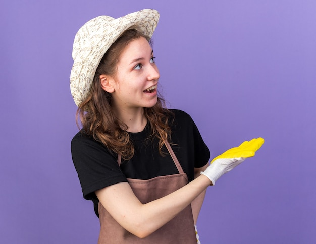 Souriant à côté jeune femme jardinier portant chapeau de jardinage avec des gants pointe sur le côté