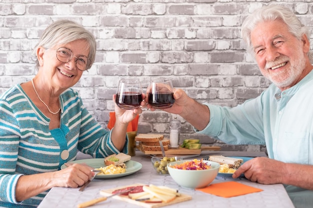 Souriant couple senior caucasien portant un toast avec un verre à vin rouge assis face à face à table en train de bruncher ensemble à la maison