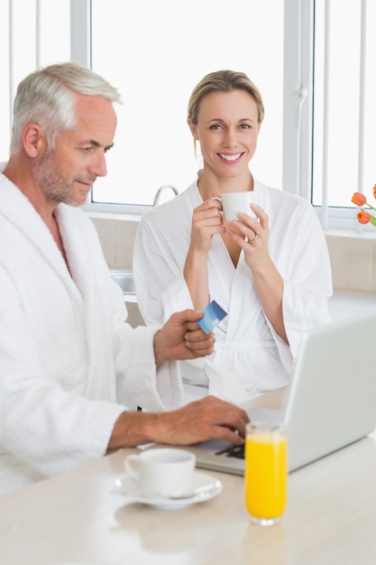 Souriant couple utilisant un ordinateur portable au petit-déjeuner dans les peignoirs