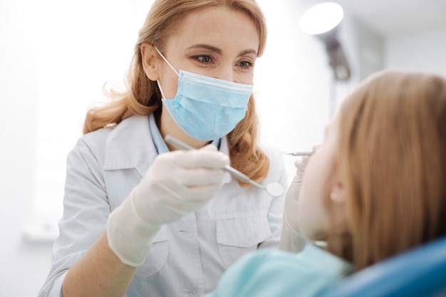 Souriant dentiste soigné compétent tenant des outils spéciaux dans ses mains et portant un uniforme tout en examinant les dents des petites filles