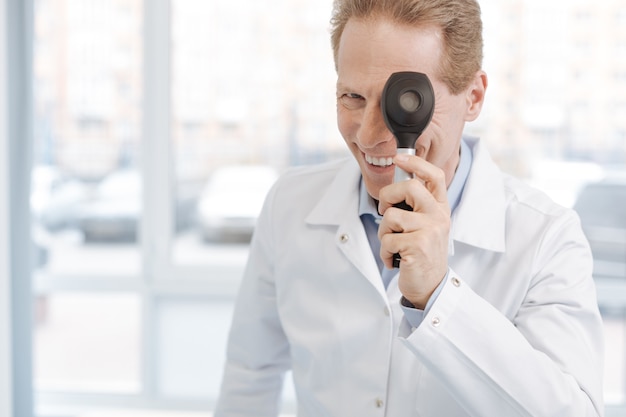 Photo souriant dermatologue positif vieillissant travaillant à l'hôpital et tenant un dermatoscope tout en exprimant sa joie