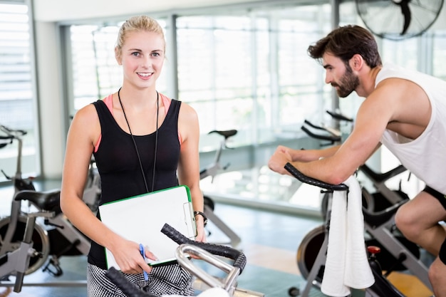 Souriant entraîneur féminin en regardant la caméra et l&#39;homme à l&#39;aide d&#39;un vélo d&#39;exercice