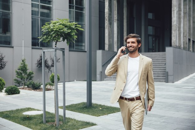 Souriant entrepreneur masculin en costume d'affaires marchant dans la rue avec un ordinateur portable et parler sur un smartphone moderne