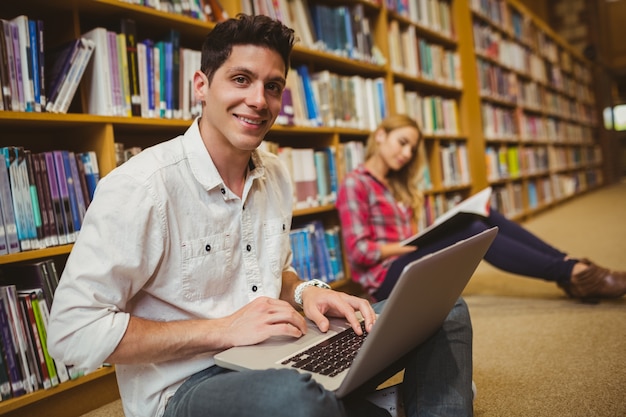 Souriant étudiant à l&#39;aide d&#39;un ordinateur portable sur le sol dans la bibliothèque