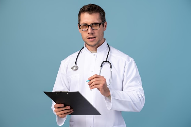 Souriant expérimenté beau jeune médecin homme isolé sur fond bleu Médecin de sexe masculin en uniforme médical stéthoscope carte de santé sur le dossier du bloc-notes Concept de médecine de santé du personnel de santé