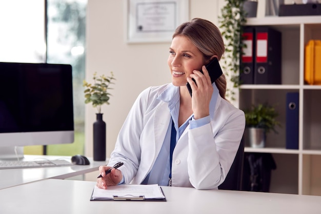 Souriant femme médecin ou médecin généraliste portant un manteau blanc assis au bureau au bureau faisant appel sur téléphone mobile