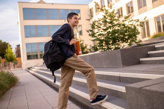 Souriant heureux adolescent aux cheveux noirs avec le sac à dos sur l'épaule en montant les escaliers en béton