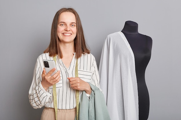 Souriant heureux joyeux égout féminin professionnel avec ruban à mesurer sur son cou à l'aide d'un téléphone portable regardant la caméra debout isolé sur fond gris