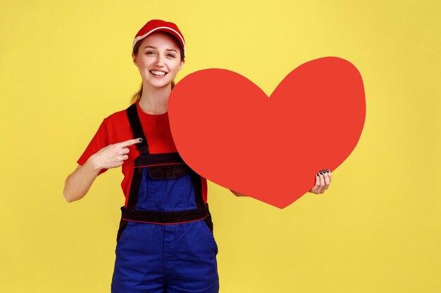 Souriant heureux travailleur femme pointant vers grand coeur rouge avec le doigt regardant la caméra exprimant des émotions positives portant une salopette et une casquette rouge Prise de vue en studio intérieur isolé sur fond jaune