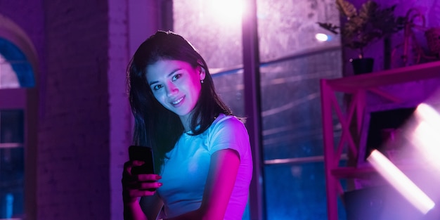 Souriant, impatient. Portrait cinématographique d'une femme élégante dans un intérieur éclairé au néon. Tonifié comme des effets de cinéma en violet-bleu. Modèle féminin caucasien utilisant un smartphone dans des lumières colorées à l'intérieur.