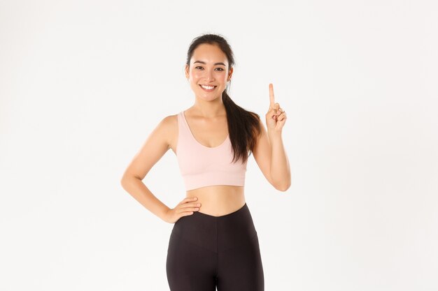 Souriant instructeur de fitness asiatique, fille en cours d'enseignement de vêtements de sport, montrant la règle numéro un, lever le doigt et sourire narquois, debout fond blanc.