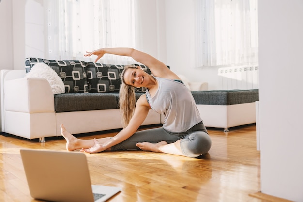 Souriant instructeur de fitness musculaire assis à la maison sur le sol et montrant des exercices d'étirement. Elle suit des cours en ligne sur un ordinateur portable.