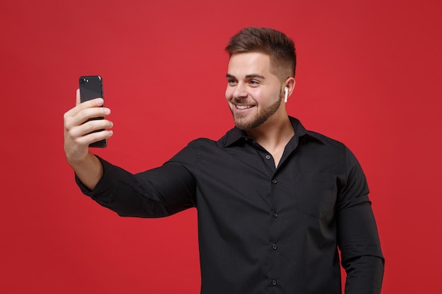 Souriant jeune barbu en chemise noire classique posant isolé sur fond rouge. Concept de style de vie des gens. Maquette de l'espace de copie. Utiliser des airpods pour faire des selfies ou passer des appels vidéo sur un téléphone portable.