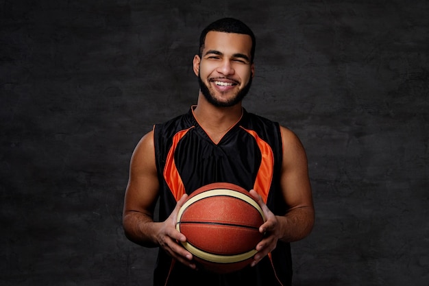 Souriant jeune basketteur afro-américain en vêtements de sport isolé sur fond sombre.