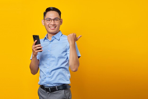 Souriant jeune bel homme d'affaires en chemise bleue classique à l'aide de smartphone et pointant avec les doigts de côté sur l'espace de copie maquette isolé sur fond jaune Atteindre le concept d'entreprise de richesse de carrière