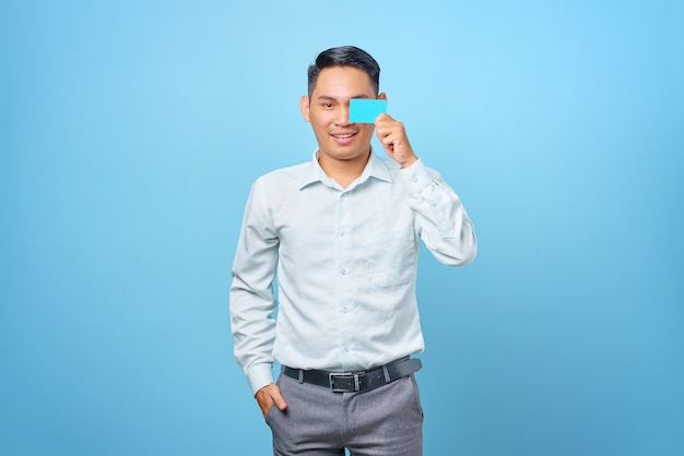 Souriant jeune bel homme d'affaires couvrant les yeux avec une carte de crédit sur fond bleu