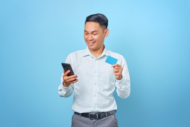 Souriant jeune bel homme d'affaires tenant un smartphone et une carte de crédit sur fond bleu