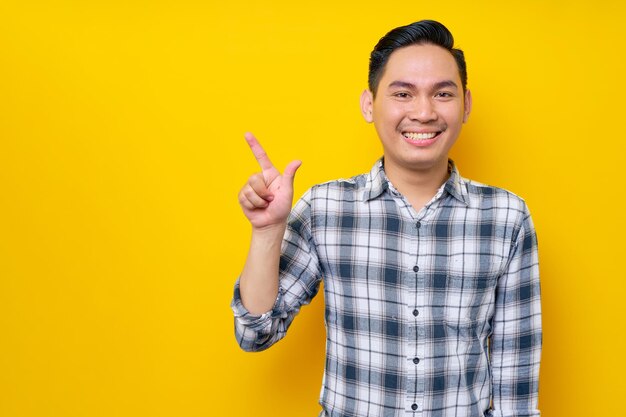 Souriant jeune bel homme asiatique portant une chemise à carreaux pointant du doigt l'espace de copie vide isolé sur fond jaune Concept de style de vie des gens