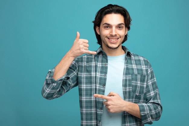 souriant jeune bel homme montrant un geste d'appel regardant la caméra pointant vers le côté isolé sur fond bleu