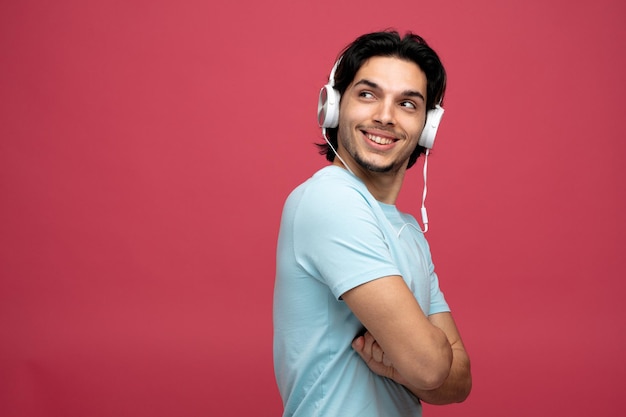 souriant jeune bel homme portant des écouteurs debout dans la vue de profil avec les bras croisés regardant le côté isolé sur fond rouge avec espace de copie