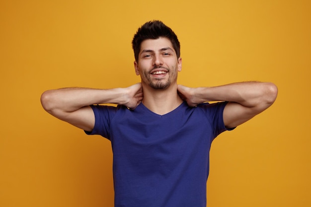 Souriant jeune bel homme regardant la caméra en gardant les mains derrière le cou sur fond jaune