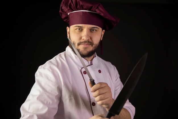 Souriant jeune chef barbu en uniforme tient des couteaux tranchants croisés