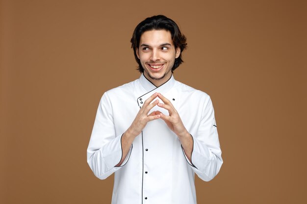 souriant jeune chef masculin portant l'uniforme gardant les mains ensemble regardant le côté isolé sur fond marron