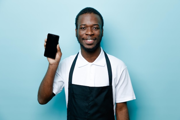 Souriant jeune coiffeur afro-américain en uniforme tenant le téléphone isolé sur fond bleu