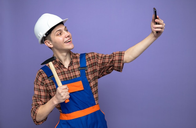 Souriant jeune constructeur masculin en uniforme avec marteau prendre un selfie