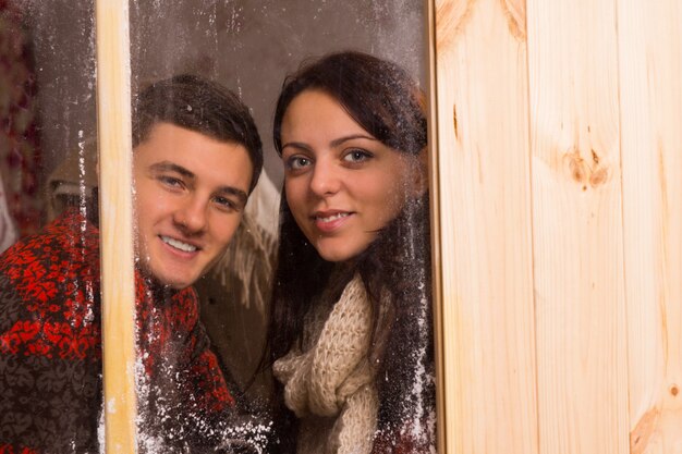 Souriant jeune couple debout côte à côte regardant à travers une vitre givrée en hiver et regardant la caméra avec un sourire