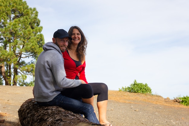 Souriant jeune couple posant assis sur un tronc d'arbre à la forêt par une journée ensoleillée. Homme sur capot et casquette et femme reposant joyeusement sur trek de randonnée