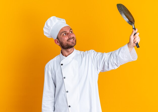 Souriant jeune cuisinier masculin caucasien en uniforme de chef et casquette levant et regardant une poêle à frire isolée sur un mur orange