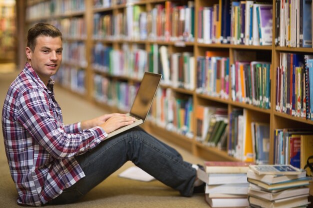 Souriant jeune étudiant assis sur le sol de la bibliothèque à l&#39;aide d&#39;un ordinateur portable