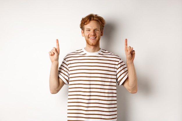 Souriant jeune étudiant aux cheveux rouges montrant le logo, pointant les doigts vers le haut et l'air heureux, debout sur fond blanc.