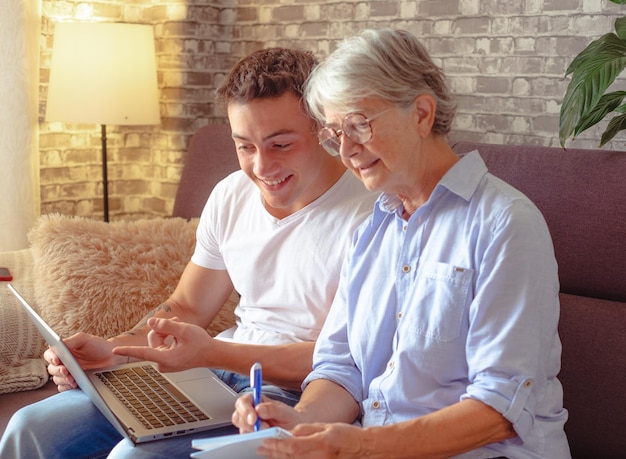 Souriant jeune garçon assis sur un canapé à la maison aidant une grand-mère aînée à surfer avec un ordinateur portable sur le Web pendant qu'elle prend des notes