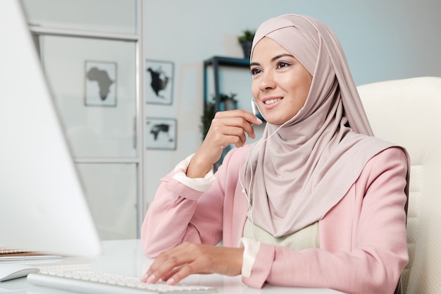 Souriant jeune gestionnaire musulman en hijab rose à l'aide de pièces d'oreille tout en communiquant via l'application de vidéoconférence sur ordinateur au bureau