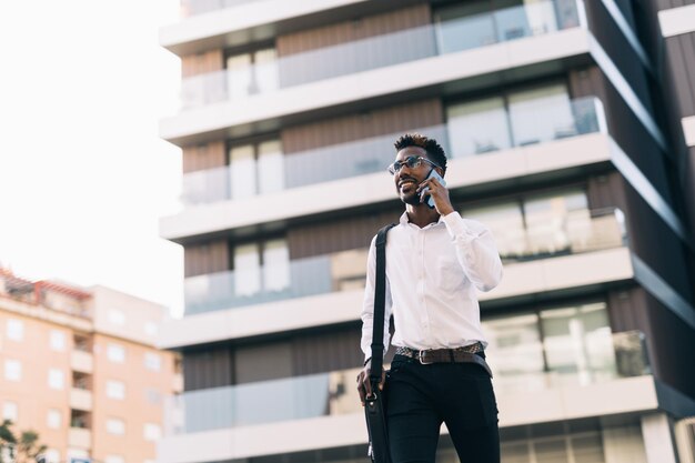 Souriant jeune homme d'affaires afro-américain marchant dans la rue en parlant sur son téléphone intelligent